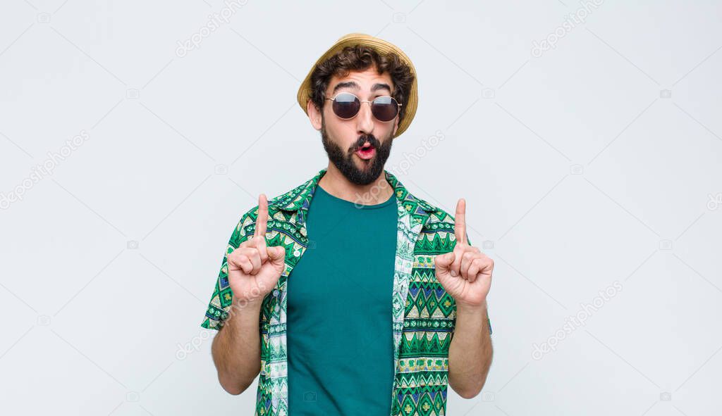 young tourist man feeling awed and open mouthed pointing upwards with a shocked and surprised look against white wall