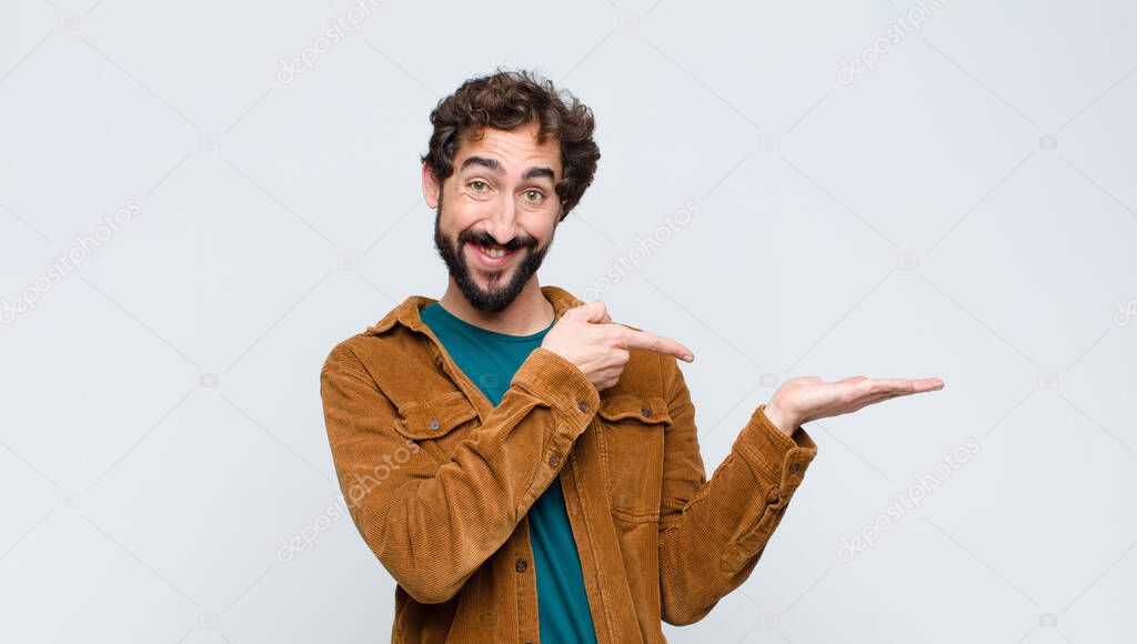 young handsome man smiling cheerfully and pointing to copy space on palm on the side, showing or advertising an object against flat wall