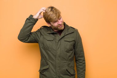 Genç sarışın adam şaşkın ve şaşkın hissediyor, başını kaşıyor ve düz duvara bakıyor.