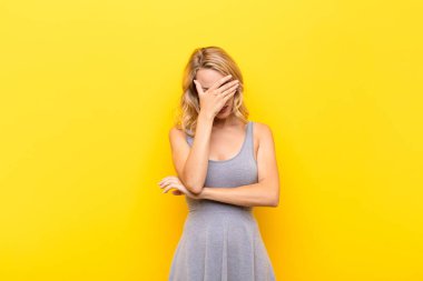 Genç sarışın kadın stresli, utanmış ya da üzgün görünüyor, baş ağrısı var, yüzünü turuncu duvara dayamış.