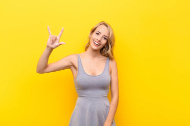 Genç sarışın kadın mutlu, eğlenceli, kendinden emin, pozitif ve isyankar hissediyor, elleri turuncu duvara dayalı kaya veya heavy metal işareti yapıyor.