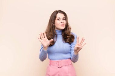 Genç, güzel bir bayan gülümsüyor ve arkadaş canlısı görünüyor. 9 ya da 9 numarayı gösteriyor. Elleri önde, bej arka planda geriye doğru sayıyor.