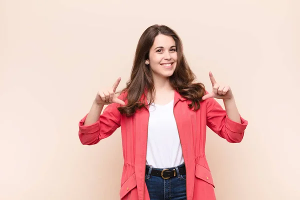 young pretty woman framing or outlining own smile with both hands, looking positive and happy, wellness concept against beige background