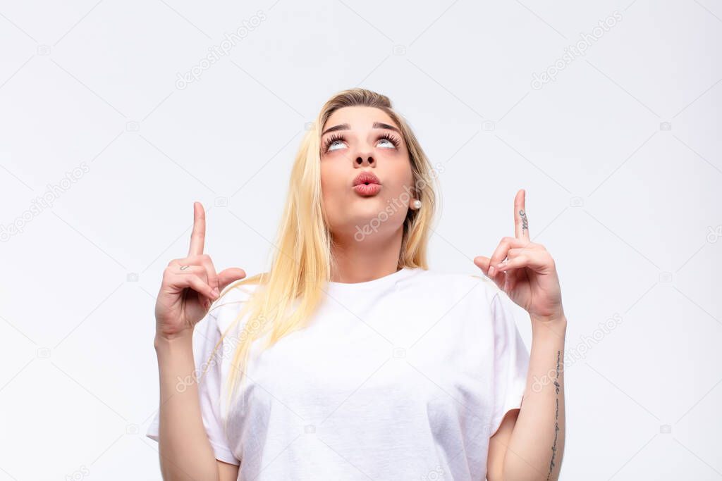 young pretty blonde woman looking shocked, amazed and open mouthed, pointing upwards with both hands to copy space against white wall
