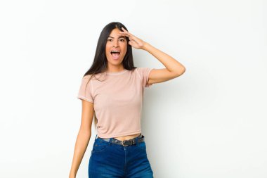 Genç ve güzel Latin kadın mutlu, şaşkın ve şaşırmış görünüyor, gülümsüyor ve düz duvara karşı inanılmaz ve inanılmaz iyi haberler veriyor.