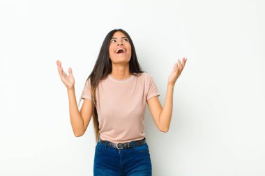 Genç, güzel, Latin bir kadın kendini mutlu, şaşkın, şanslı ve şaşırmış hissediyor. Zaferi kutlarken iki eli de havada, düz duvara karşı.