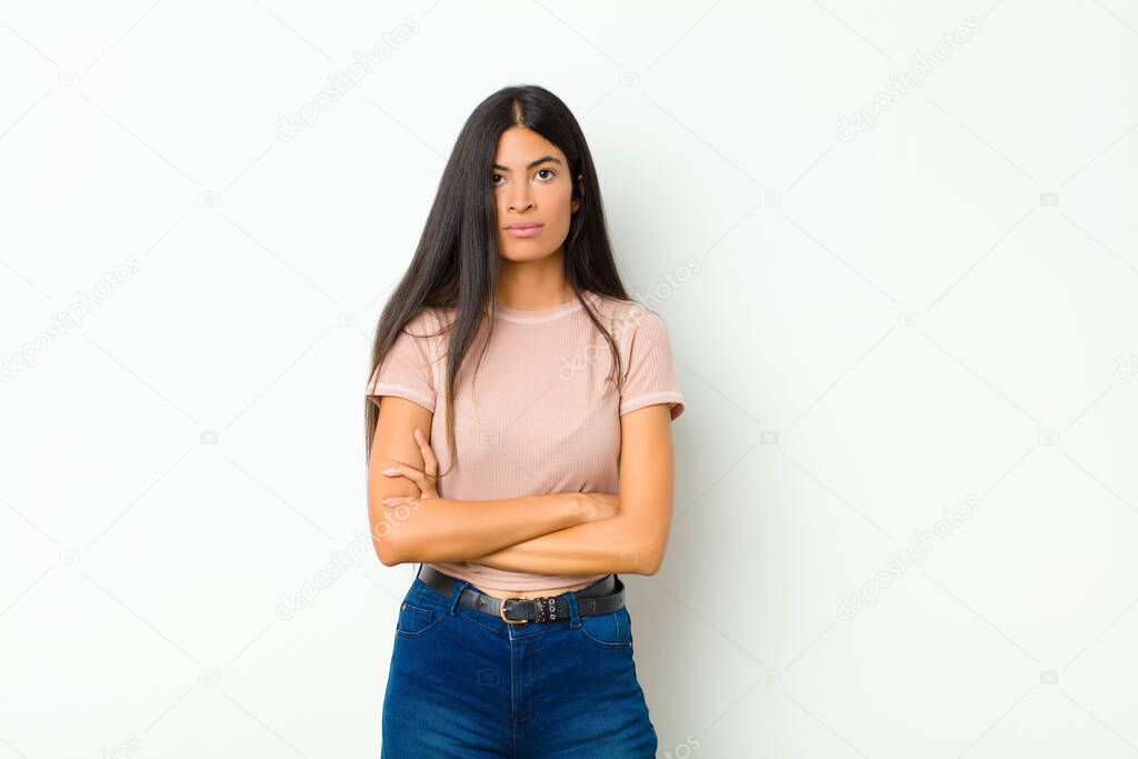 young pretty latin woman feeling displeased and disappointed, looking serious, annoyed and angry with crossed arms against flat wall