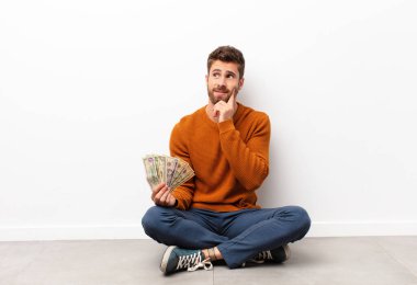 Genç yakışıklı adam mutlu bir şekilde gülümsüyor ve hayal kuruyor ya da şüpheye düşüyor, elinde dolar banknotları tutuyor.