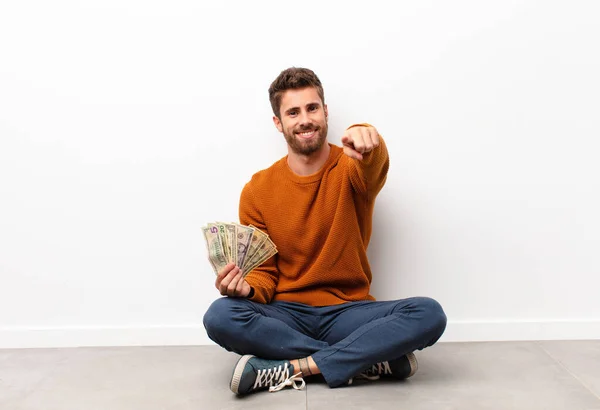 young handsome man pointing at camera with a satisfied, confident, friendly smile, choosing you holding dollar banknotes
