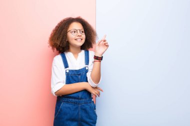 Afro-Amerikan küçük kızı mutlu bir şekilde gülümsüyor ve yan bakıyor, merak ediyor, düşünüyor ya da düz duvara karşı bir fikri var.