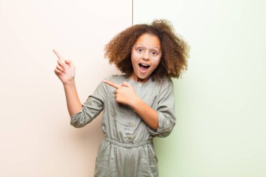 Afro-Amerikan küçük kızı kendini neşeli ve şaşırmış hissediyor, şok olmuş bir ifadeyle gülümsüyor ve yassı duvara doğru yan tarafı işaret ediyor.