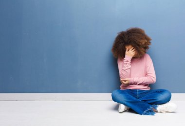 Afro-amerikan küçük kızı stresli, utanmış ya da üzgün görünüyor, baş ağrısı var, yüzü örtülü, yerde oturuyor.