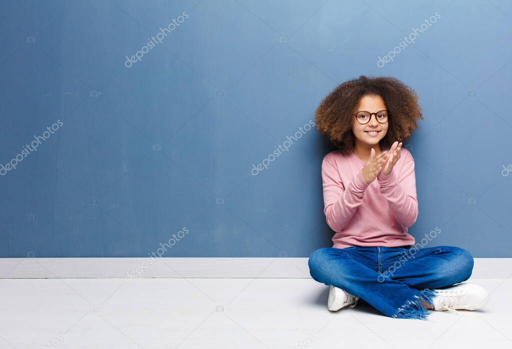 african american little girl feeling happy and successful, smiling and clapping hands, saying congratulations with an applause sitting on the floor