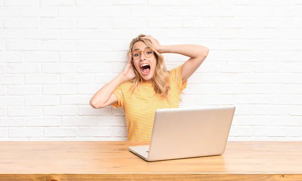 young blonde woman raising hands to head, open-mouthed, feeling extremely lucky, surprised, excited and happy using a laptop