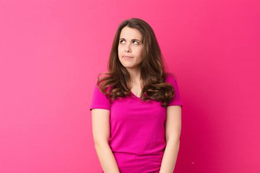 Genç ve güzel bir kadın şaşkın ve şaşkın görünüyor, merak ediyor veya bir sorunu çözmeye çalışıyor veya pembe duvara karşı düşünüyor.