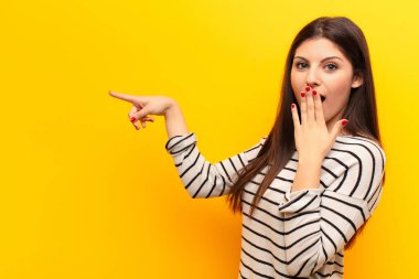 Genç, güzel bir kadın kendini mutlu, şok olmuş ve şaşırmış hissediyor. Ağzını eliyle kapatıyor ve sarı duvara karşı yanal kopya alanını işaret ediyor.