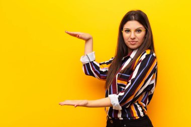 Genç ve güzel bir kadın, iki eli de kenarda olan bir objeyi tutuyor. Fotokopi alanı, sarı duvara karşı bir objeyi gösteriyor, sunuyor ya da reklamını yapıyor.