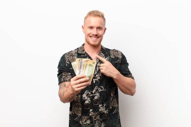 Genç sarışın adam neşeyle gülümsüyor, mutlu hissediyor ve kenarı ve yukarıyı işaret ediyor, fotokopi alanındaki objeleri banknotlarla beyaz duvara gösteriyor.