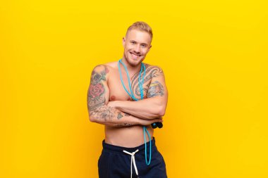 Genç ve güçlü sarışın adam kollarını kavuşturup mutlu bir şekilde gülüyor. Rahatlamış, pozitif ve tatmin olmuş bir poz veriyor ve ip atlıyor.