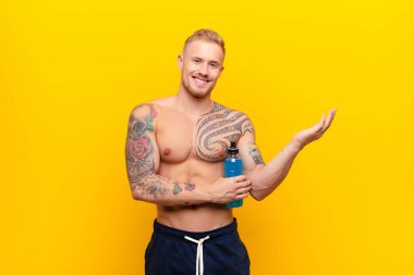 Genç sarışın güçlü adam gururla ve kendinden emin bir şekilde gülümsüyor, mutlu ve tatmin olmuş hissediyor ve fotokopi alanında enerji içeceğiyle bir konsept gösteriyor.