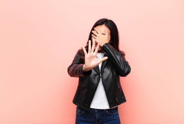 Genç, Latin, güzel bir kadın elini yüzünü örtüyor ve diğer elini kamerayı durdurmak için öne koyuyor. Duvara karşı fotoğraf veya resim çekmeyi reddediyor.