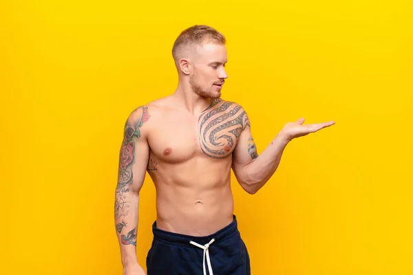 young strong blonde man feeling happy and smiling casually, looking to an object or concept held on the hand on the side against yellow wall