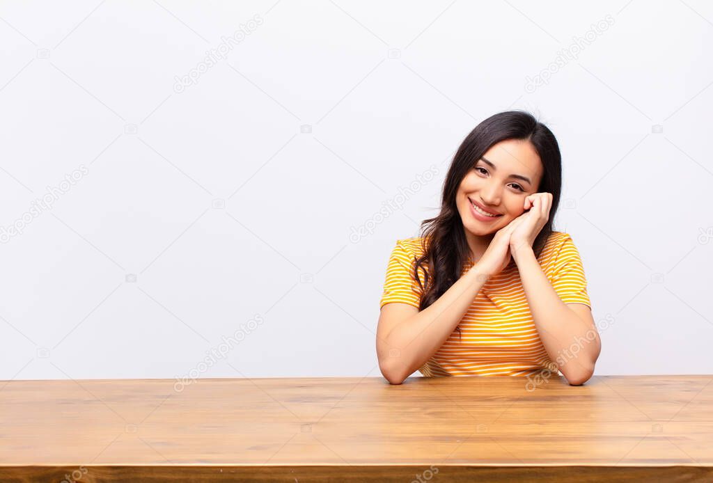 young latin pretty woman feeling in love and looking cute, adorable and happy, smiling romantically with hands next to face against flat wall