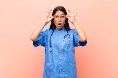 Genç Latin hemşire şaşkın, şaşkın ve şaşırmış hissediyor. Elinde pembe duvara karşı hayret dolu, inançsız bir bakış olan gözlükler tutuyor.