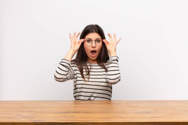 young pretty woman feeling shocked, amazed and surprised, holding glasses with astonished, disbelieving look sitting at a table clipart