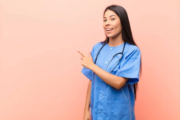 young latin nurse looking excited and surprised pointing to the side and upwards to copy space against pink wall