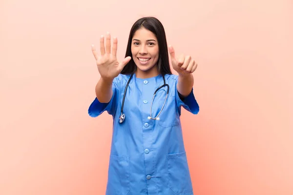 young latin nurse smiling and looking friendly, showing number six or sixth with hand forward, counting down against pink wall