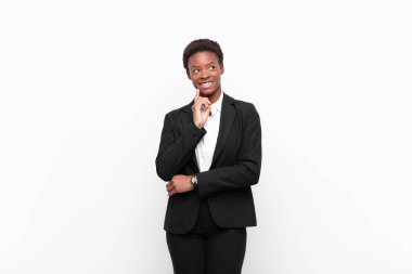 Genç ve güzel siyah kadın mutlu bir şekilde gülümsüyor ve hayal kuruyor ya da şüphe ediyor, beyaz duvara bakıyor.