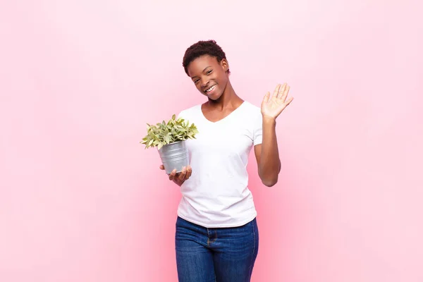 young pretty black womansmiling happily and cheerfully, waving hand, welcoming and greeting you, or saying goodbye holding a plant