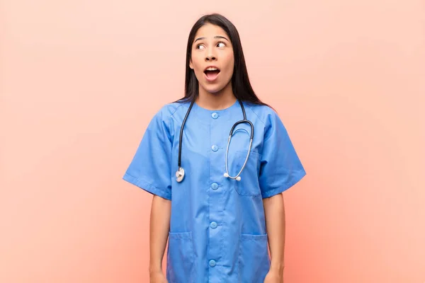 young latin nurse feeling shocked, happy, amazed and surprised, looking to the side with open mouth against pink wall