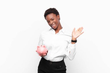 young pretty black womansmiling happily and cheerfully, waving hand, welcoming and greeting you, or saying goodbye holding a piggy bank clipart