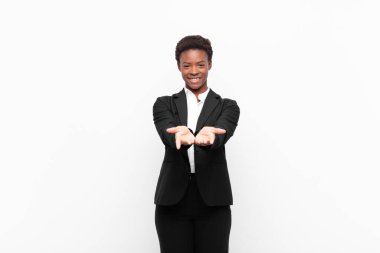 Genç, güzel siyah kadın mutlu bir şekilde gülümsüyor. Dost canlısı, kendine güvenen, pozitif bakışlı, beyaz duvara karşı bir nesne ya da konsept sunuyor ve gösteriyor.