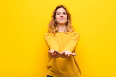 Genç, güzel sarışın kadın mutlu bir şekilde gülümsüyor. Cana yakın, kendinden emin, pozitif bakışlı. Sarı duvara karşı bir nesne ya da konsept sunuyor ve gösteriyor.