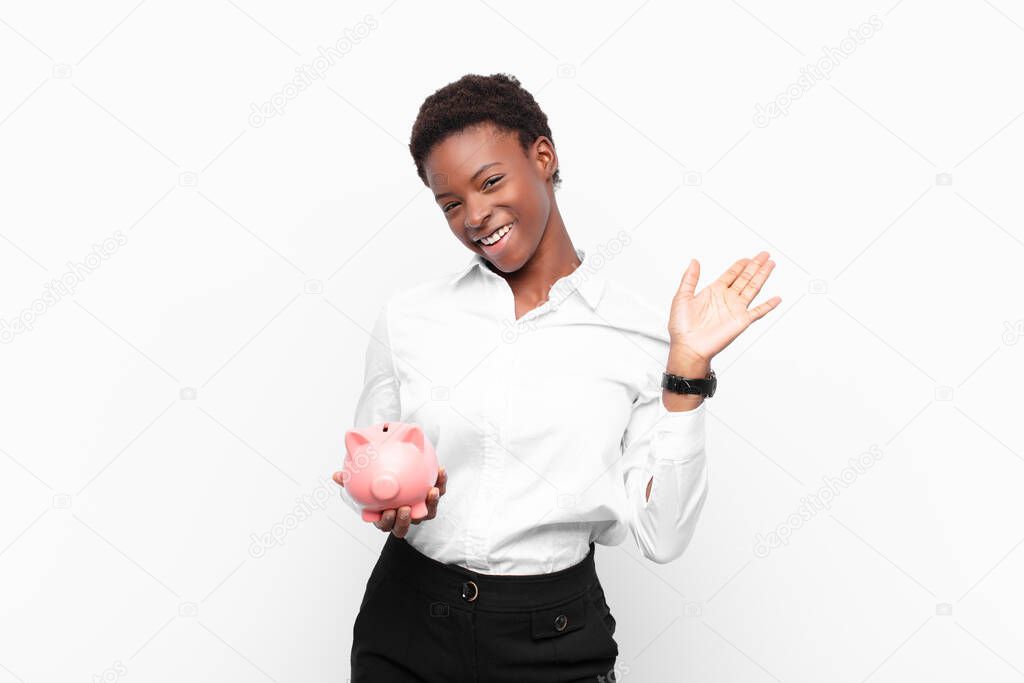 young pretty black womansmiling happily and cheerfully, waving hand, welcoming and greeting you, or saying goodbye holding a piggy bank