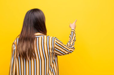 Genç ve güzel bir kadın dikilip fotokopi alanındaki objeyi işaret ediyor, turuncu duvara karşı dikiz manzarası
