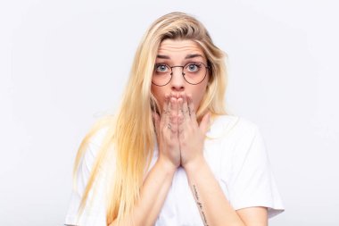 young pretty blonde woman feeling worried, upset and scared, covering mouth with hands, looking anxious and having messed up against white wall clipart