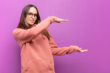 Genç, güzel bir kadın gülümsüyor, mutlu, pozitif ve tatmin olmuş hissediyor. Mor duvara karşı fotokopi alanında nesne veya konsept tutuyor veya gösteriyor.