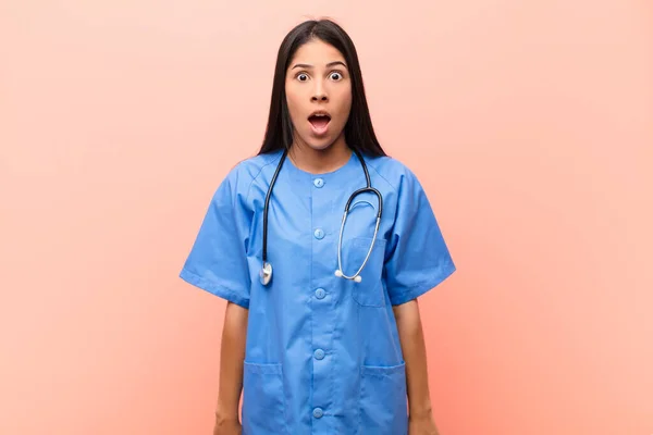 young latin nurse looking very shocked or surprised, staring with open mouth saying wow against pink wall