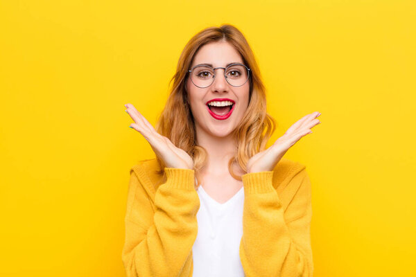 молодая красивая блондинка выглядит счастливой и взволнованной, шокированной неожиданным сюрпризом с обеими руками, открытыми рядом с лицом к желтой стене