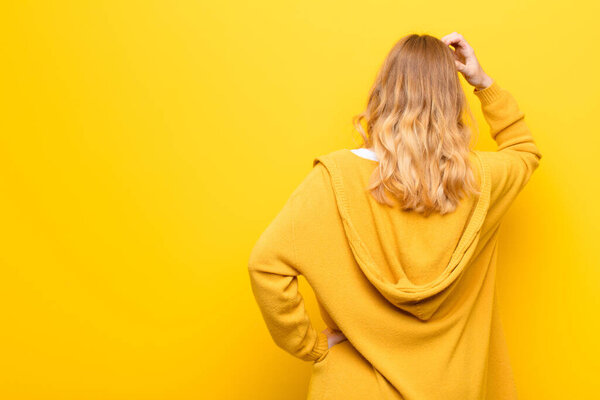 молодая красивая блондинка чувствует себя невежественной и смущенной, думая о решении, с рукой на бедре и другой на голове, вид сзади на желтую стену