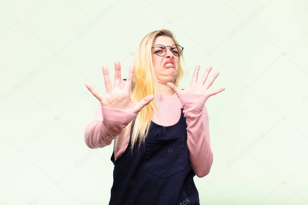 young pretty blonde woman feeling terrified, backing off and screaming in horror and panic, reacting to a nightmare against flat wall