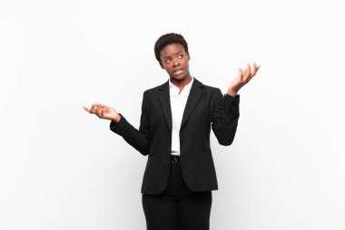 young pretty black womanshrugging with a dumb, crazy, confused, puzzled expression, feeling annoyed and clueless against white wall