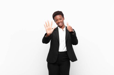 Genç ve güzel siyah kadın gülümsüyor ve arkadaş canlısı görünüyor. Altı ya da altıncı numarayı gösteriyor. Elleri önde, beyaz duvara doğru sayıyor.