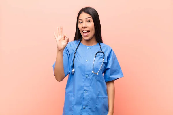 young latin nurse feeling successful and satisfied, smiling with mouth wide open, making okay sign with hand against pink wall