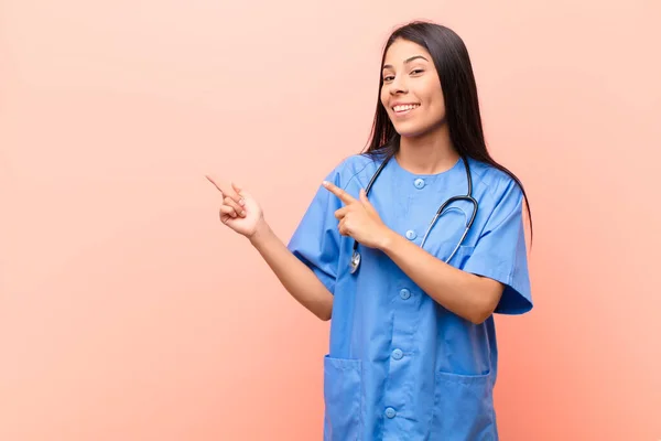 young latin nurse smiling happily and pointing to side and upwards with both hands showing object in copy space against pink wall