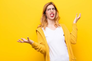 young pretty blonde woman shrugging with a dumb, crazy, confused, puzzled expression, feeling annoyed and clueless against yellow wall
